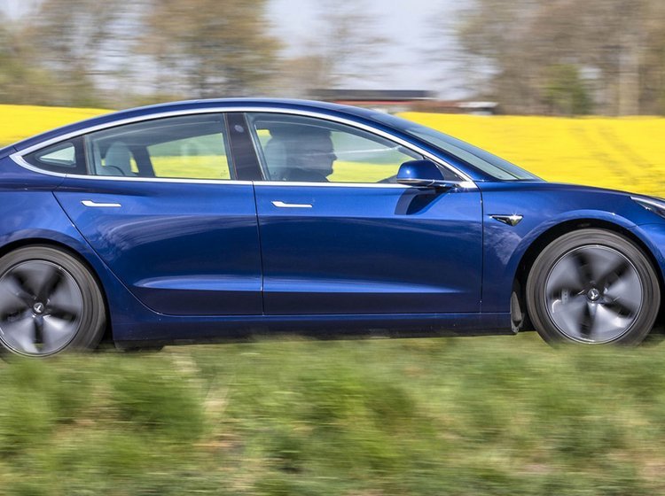 En blå Tesla Model 3 kører forbi en gul rapsmark. 