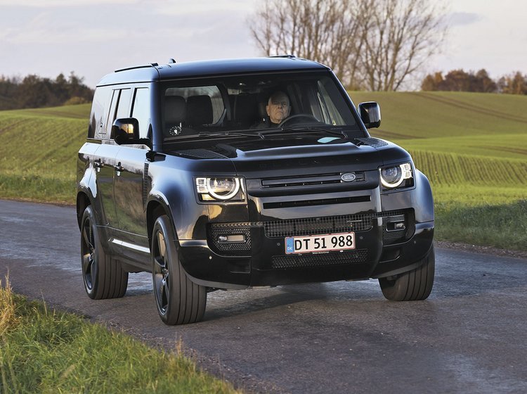 Land Rover Defender kører på en landevej med græs i baggrunden.