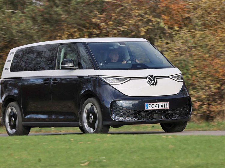 En blå og hvid Volkswagen ID. Buzz elektrisk minibus fotograferet under kørsel på en grøn græsmark med træer i baggrunden. 