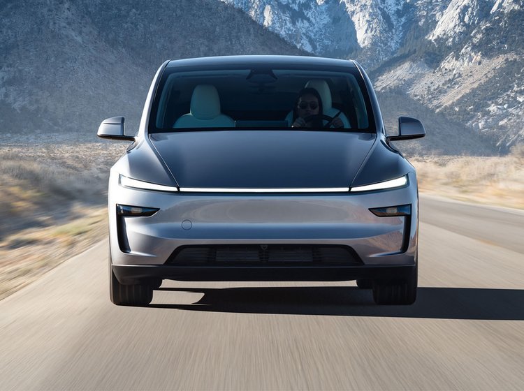 En grå faceliftet Tesla Model Y kører på vej med bjerge i baggrunden. 