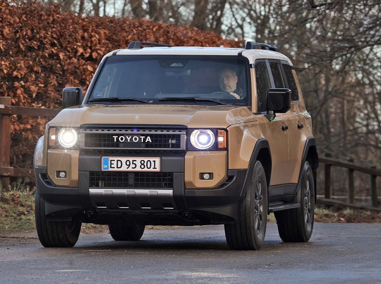 En Toyota Land Cruiser set skråt forfra kører på landevej med hegn og buske i baggrunden