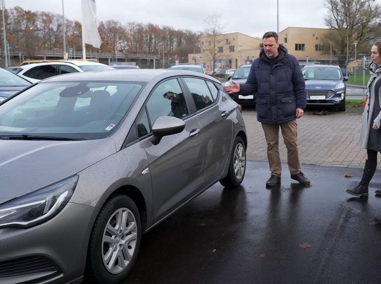 En mand peger på en grå bil. En anden mand og en dame ser på bilen.