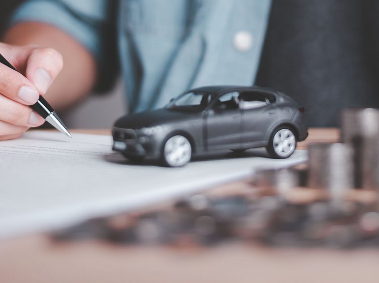 Mand sidder med kuglepen i hånden og peger på papir. På papiret ses en lille modelbil. Ved siden af en stabel af mønter. 