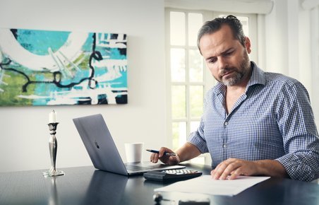 Mand sidder ved bærbar computer indenfor og kigger på papirer. 