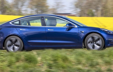 En blå Tesla Model 3 kører forbi en gul rapsmark. 