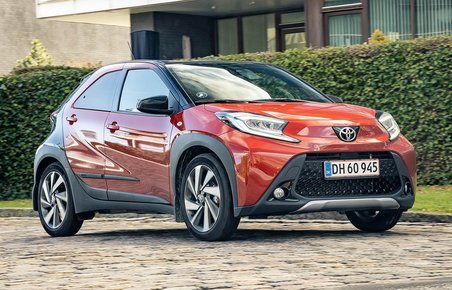 Orange Toyota Aygo X kører på brosten med grøn hæk i baggrunden.