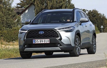 Hybridbilen Toyota Corolla Cross kører i et sving med et hvidt hus og træer i baggrunden.