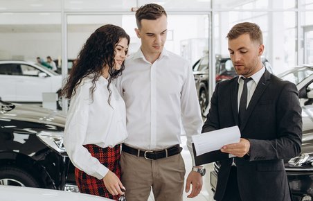 Tre personer står hos en bilforhandler og ser på et stykke papir