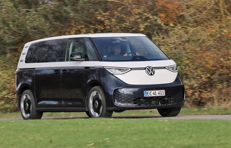 En blå og hvid Volkswagen ID. Buzz elektrisk minibus fotograferet under kørsel på en grøn græsmark med træer i baggrunden. 