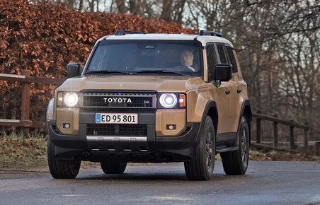 En Toyota Land Cruiser set skråt forfra kører på landevej med hegn og buske i baggrunden