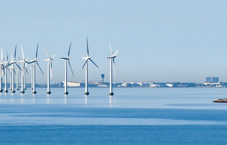 Vindmøller står på havet med kystlinjen i baggrunden.