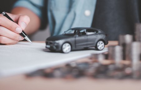 Mand sidder med kuglepen i hånden og peger på papir. På papiret ses en lille modelbil. Ved siden af en stabel af mønter. 